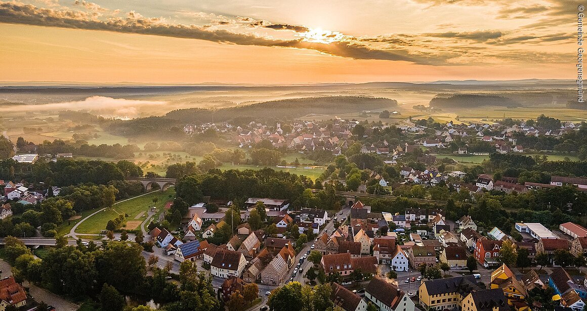 Luftaufnahme einer ländlichen Gemeinde bei Sonnenaufgang, mit Häusern, Bäumen und Feldern im Hintergrund.