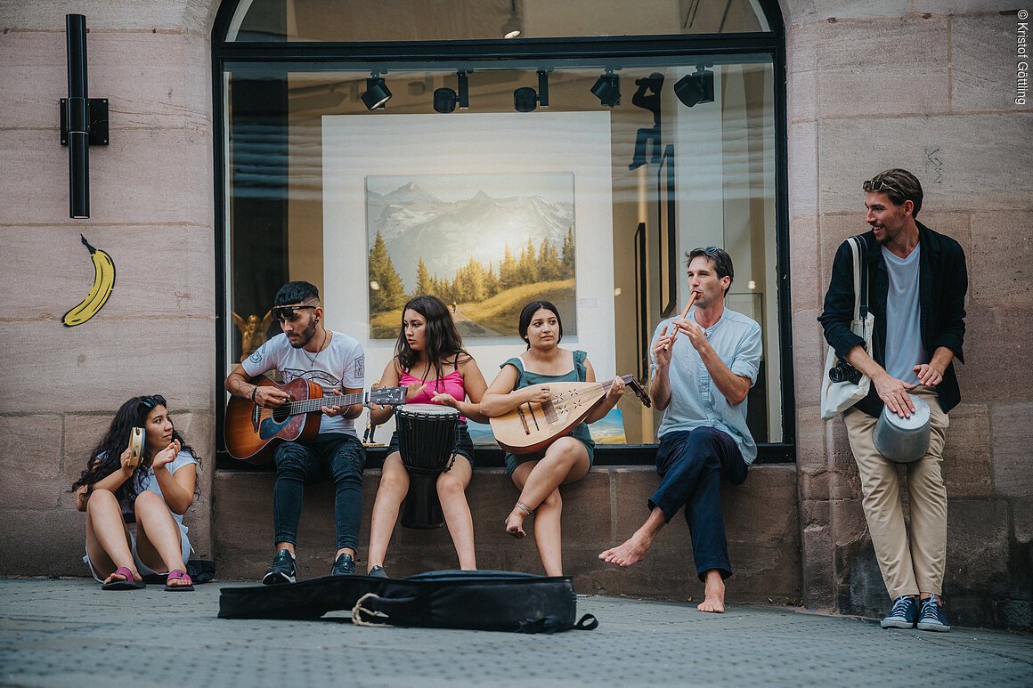Eine Gruppe von sechs Personen spielt verschiedene Musikinstrumente vor einem Schaufenster.
