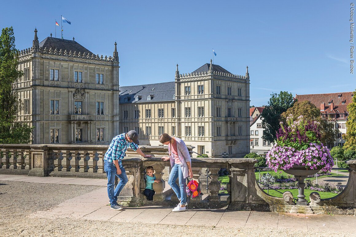 Eine Familie steht auf einer Terrasse vor einem historischen Gebäude. Ein Kind schaut durch das Geländer.