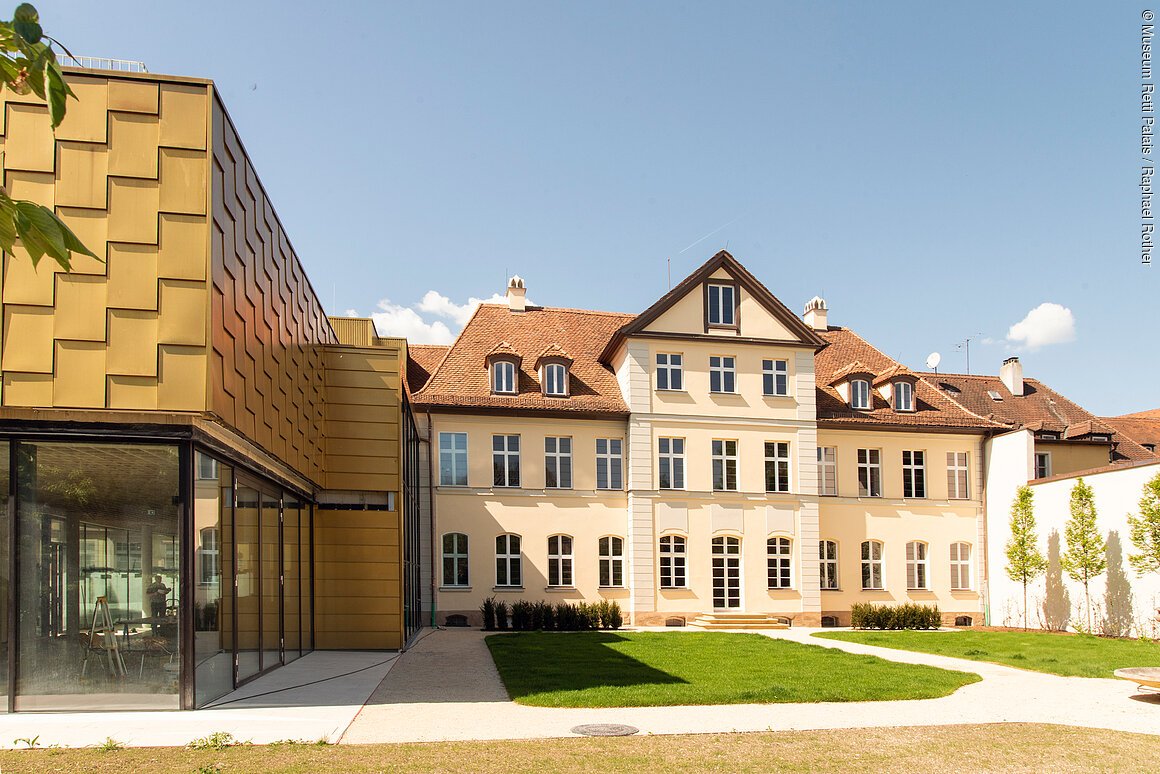 Modernes Gebäude mit goldener Fassade neben einem historischen Gebäude mit roten Ziegeldächern.