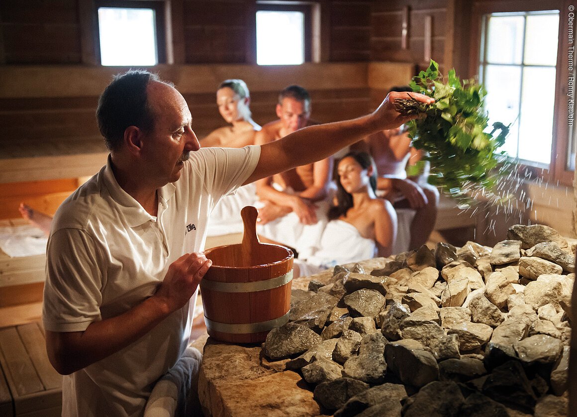 Eine Person gießt Wasser mit Kräutern über heiße Steine in einer Sauna, während andere Personen im Hintergrund sitzen.