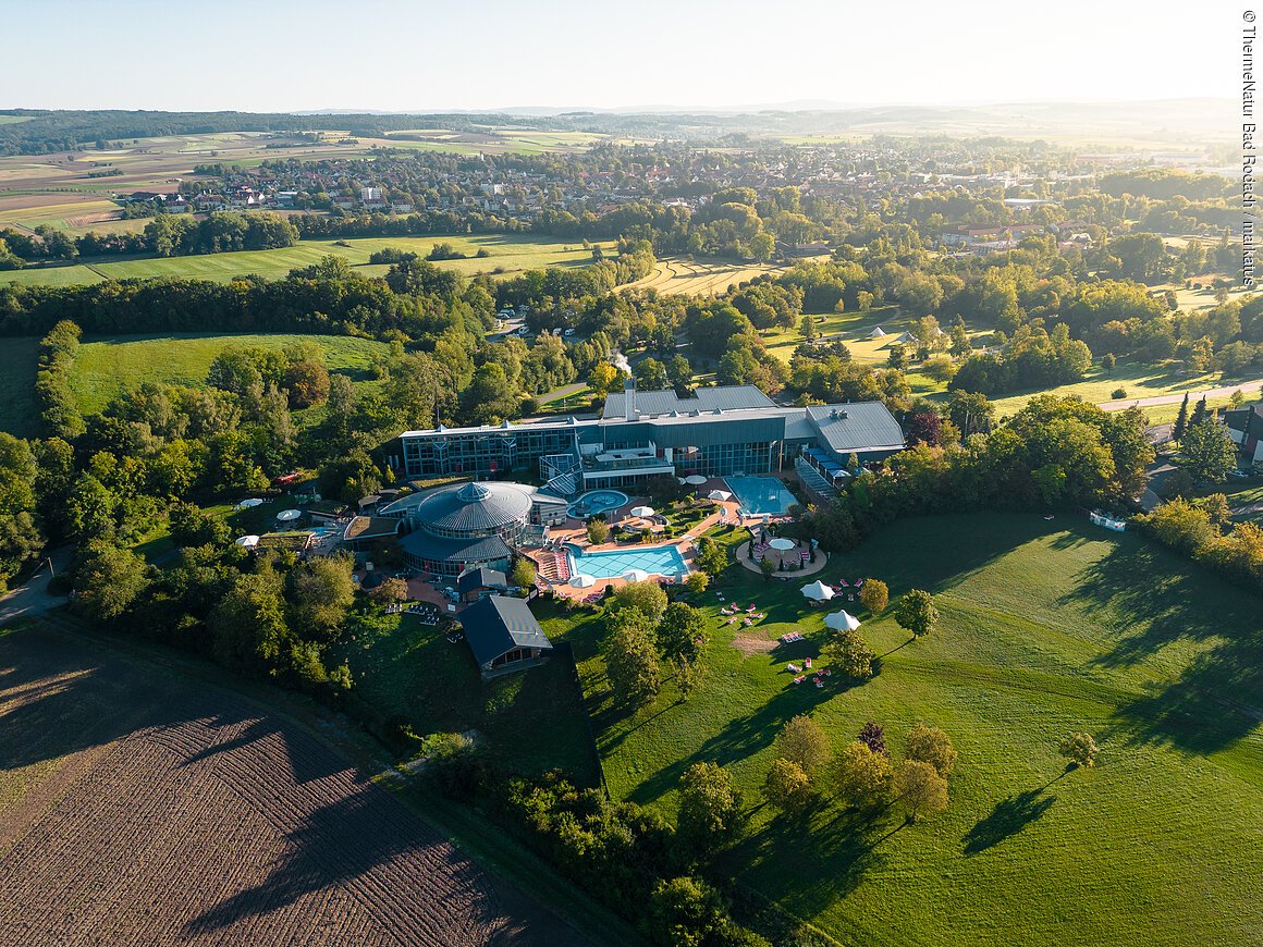Luftaufnahme eines großen Gebäudekomplexes mit Pools, umgeben von Wiesen und Bäumen, im Hintergrund eine ländliche Landschaft.
