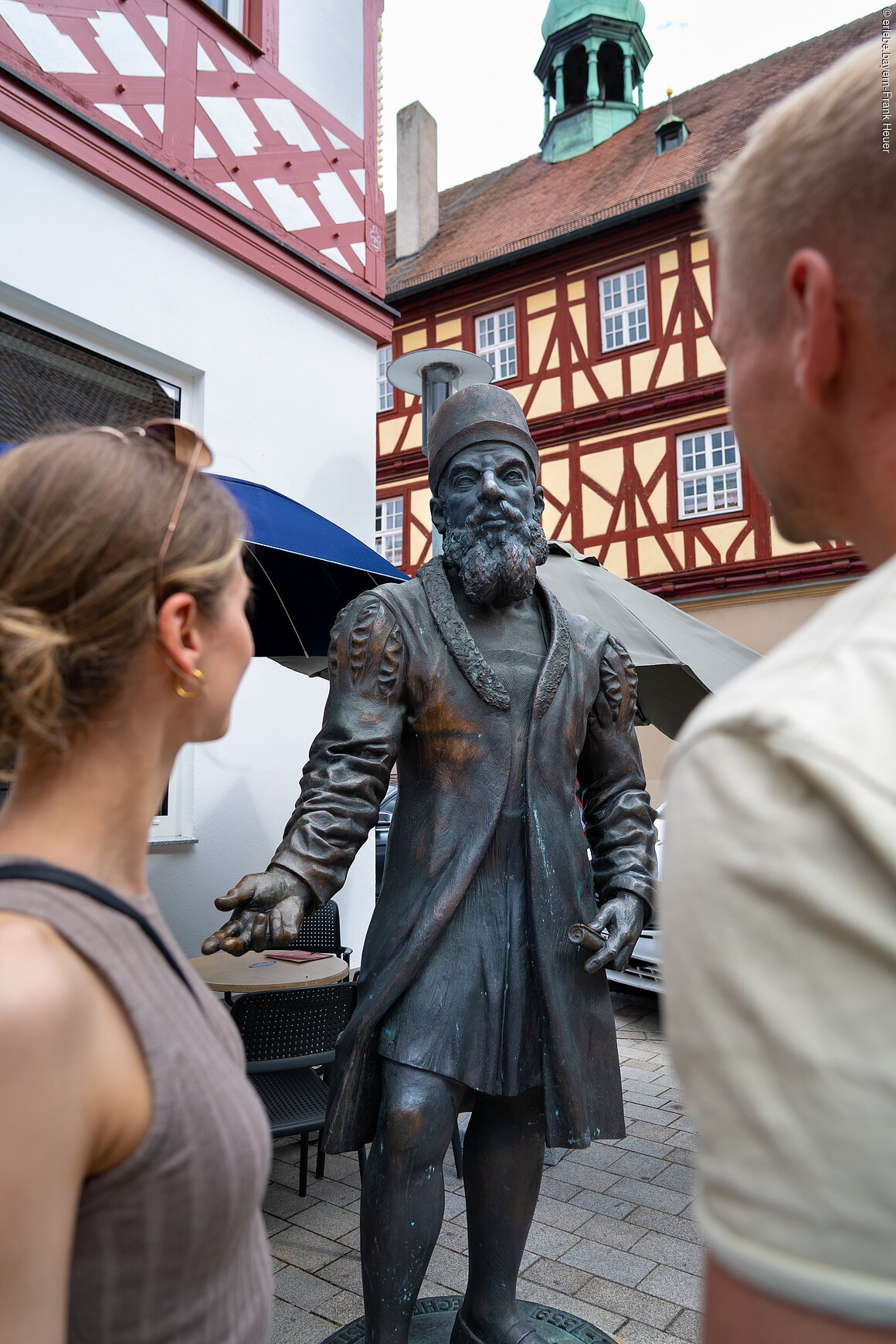 Zwei Personen betrachten eine Bronzestatue in historischer Kleidung vor Fachwerkhäusern.
