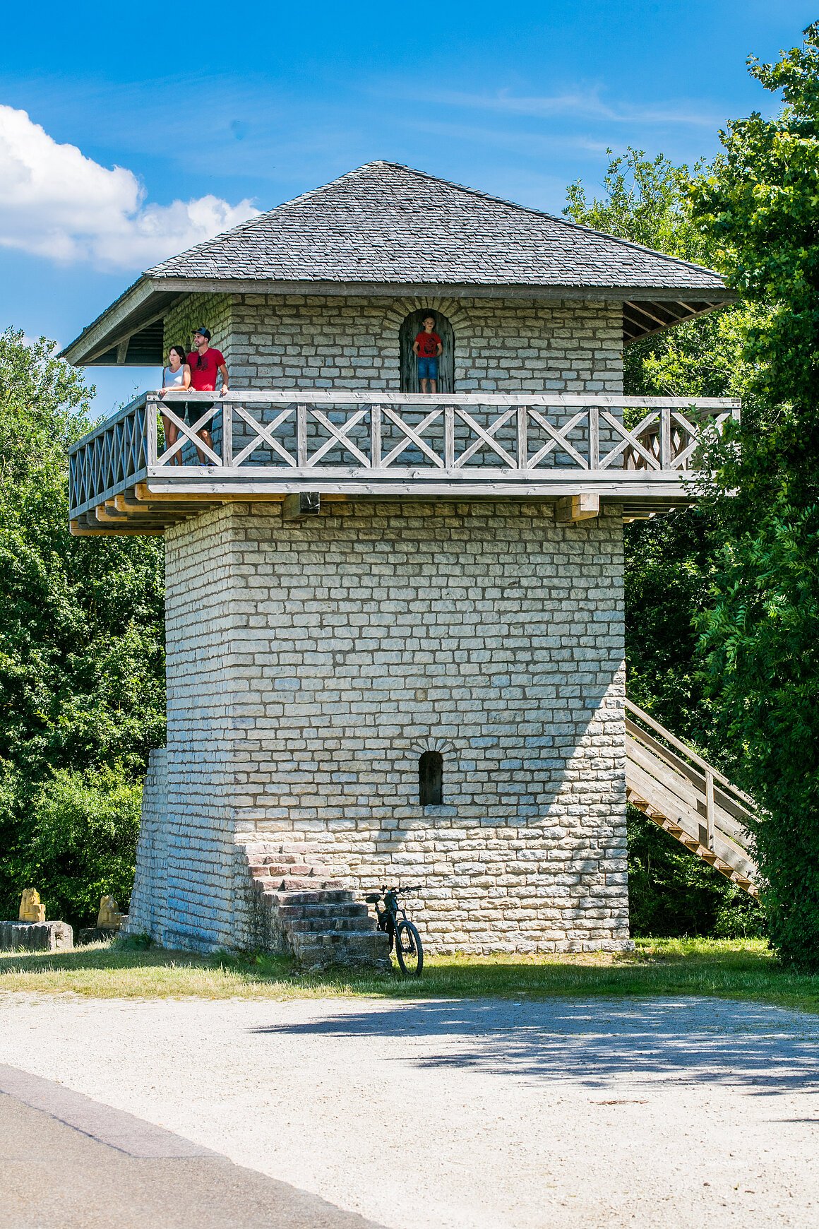 Römerturm (Titting, Naturpark Altmühltal) Steinturm mit Holzgeländer und Treppe, umgeben von Bäumen. Drei Personen auf dem Balkon. Fahrrad unten.