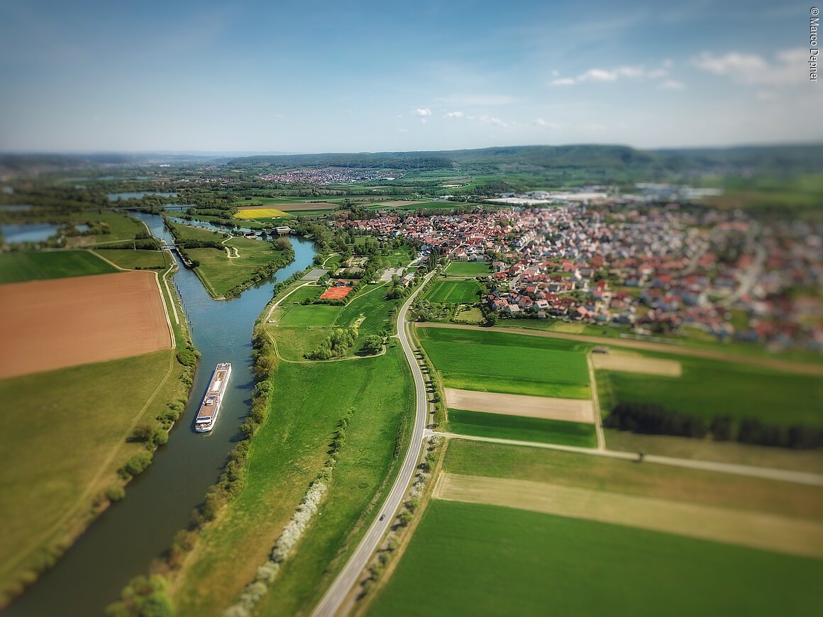 Luftaufnahme einer Flusslandschaft mit einem Frachtschiff, umgeben von Feldern und einer Siedlung im Hintergrund.
