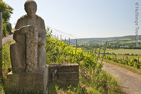 Steinerne Statue eines sitzenden Mannes mit Weintrauben in einem Weinberg, Weg und Landschaft im Hintergrund.