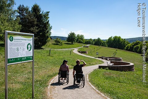 Zwei Personen im Rollstuhl auf barrierefreiem Weg mit Bänken und Infotafel im Grünen unter blauem Himmel.