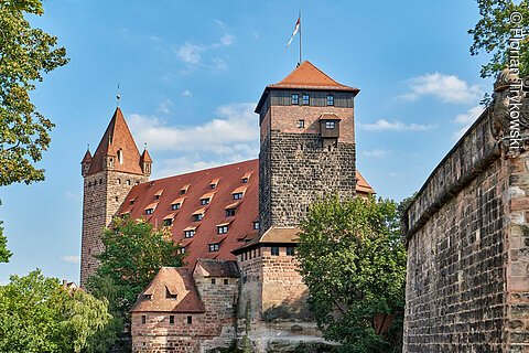 Burg mit roten Dächern und Türmen, umgeben von Bäumen und einer Steinmauer bei blauem Himmel.