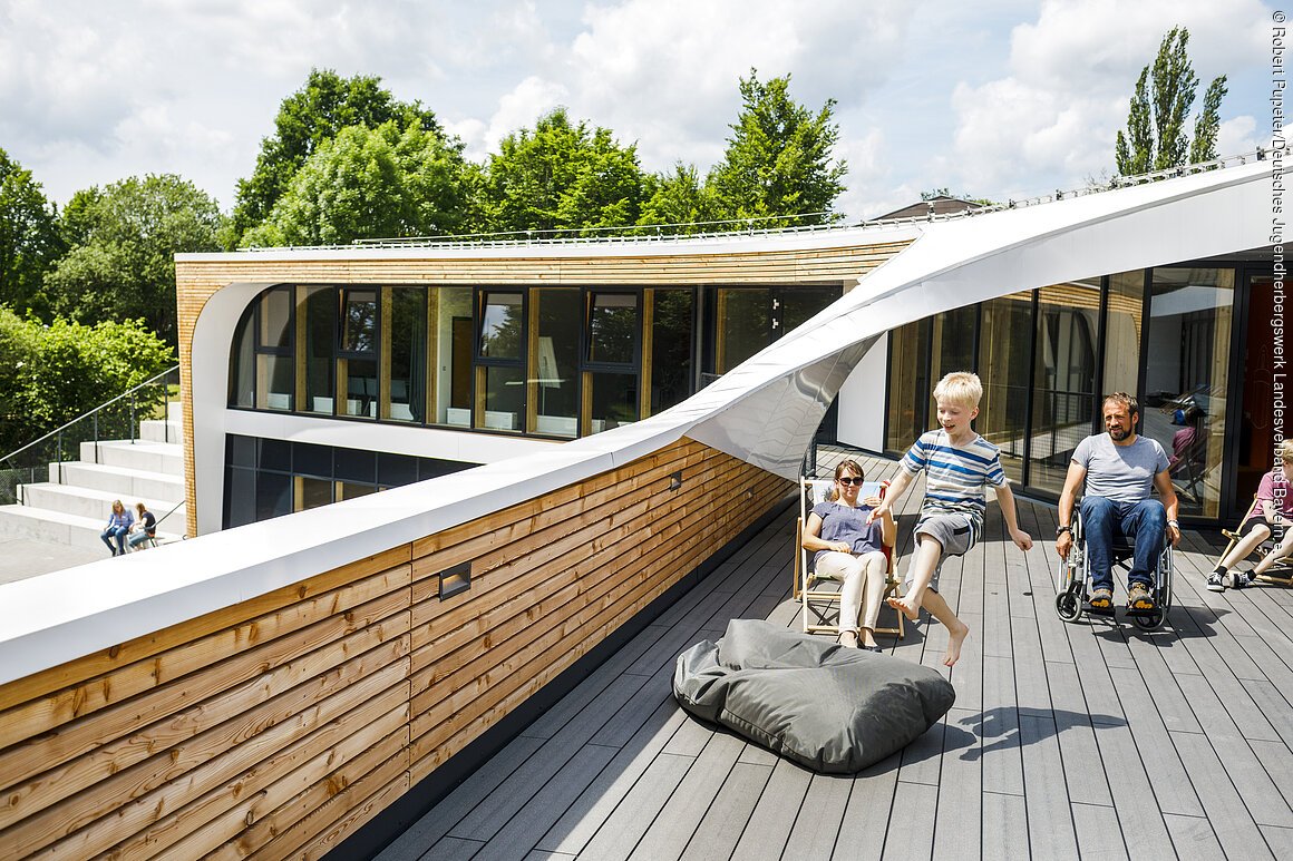 Kind springt auf Dachterrasse vor modernem Gebäude, Frau sitzt auf Liegestuhl, Mann im Rollstuhl daneben