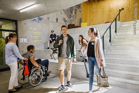 Mehrere Personen, darunter ein Mann im Rollstuhl und zwei junge Erwachsene, unterhalten sich in einem modernen Innenraum mit Betonwänden und Treppe.