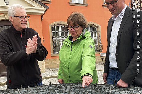 Drei Personen betrachten eine taktile Stadtmodellkarte im Freien vor einem orangefarbenen Gebäude.