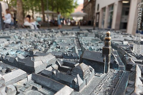 Bronzene taktile Stadtmodell mit Gebäuden und Kirche, unscharfer Hintergrund mit Menschen und Bäumen
