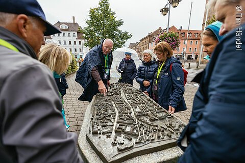 Gruppe von Menschen betrachtet bei bewölktem Wetter ein großes Stadtmodell im Freien auf einem Platz.