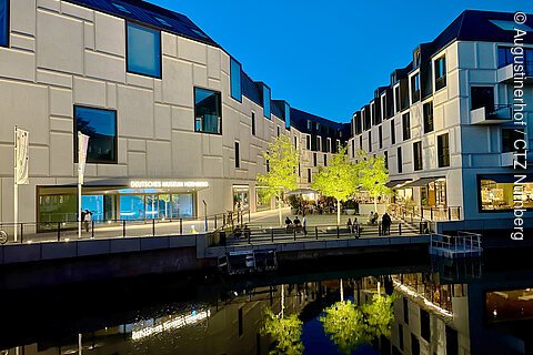 Beleuchtetes modernes Gebäude des Deutschen Museums Nürnberg mit Bäumen und Wasserreflexion bei Abenddämmerung.