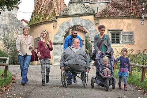 Mehrgenerationenfamilie mit Rollstuhl und Kinderwagen vor historischem Tor in Dorfstraße unterwegs