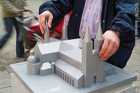 Hände berühren ein graues Architekturmodell einer Kirche mit mehreren Türmen auf einem Podest im Freien.