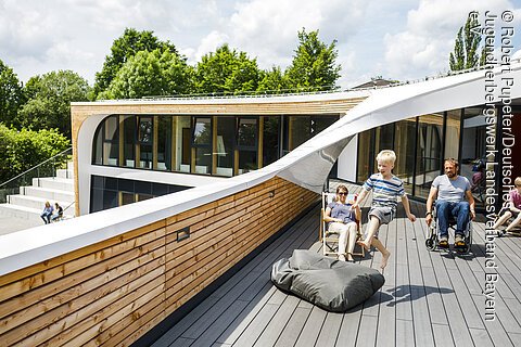 Kind springt auf Dachterrasse vor modernem Gebäude, Frau sitzt auf Liegestuhl, Mann im Rollstuhl daneben