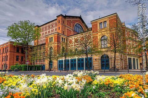 Backsteingebäude mit großen Fenstern, umgeben von blühenden Blumenbeeten und Bäumen, unter bewölktem Himmel.