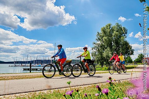 Eine Gruppe von Radfahrer:innen fährt bei schönem Wetter vor einem Gewässer über einen asphaltierten Radweg zwischen Bäumen