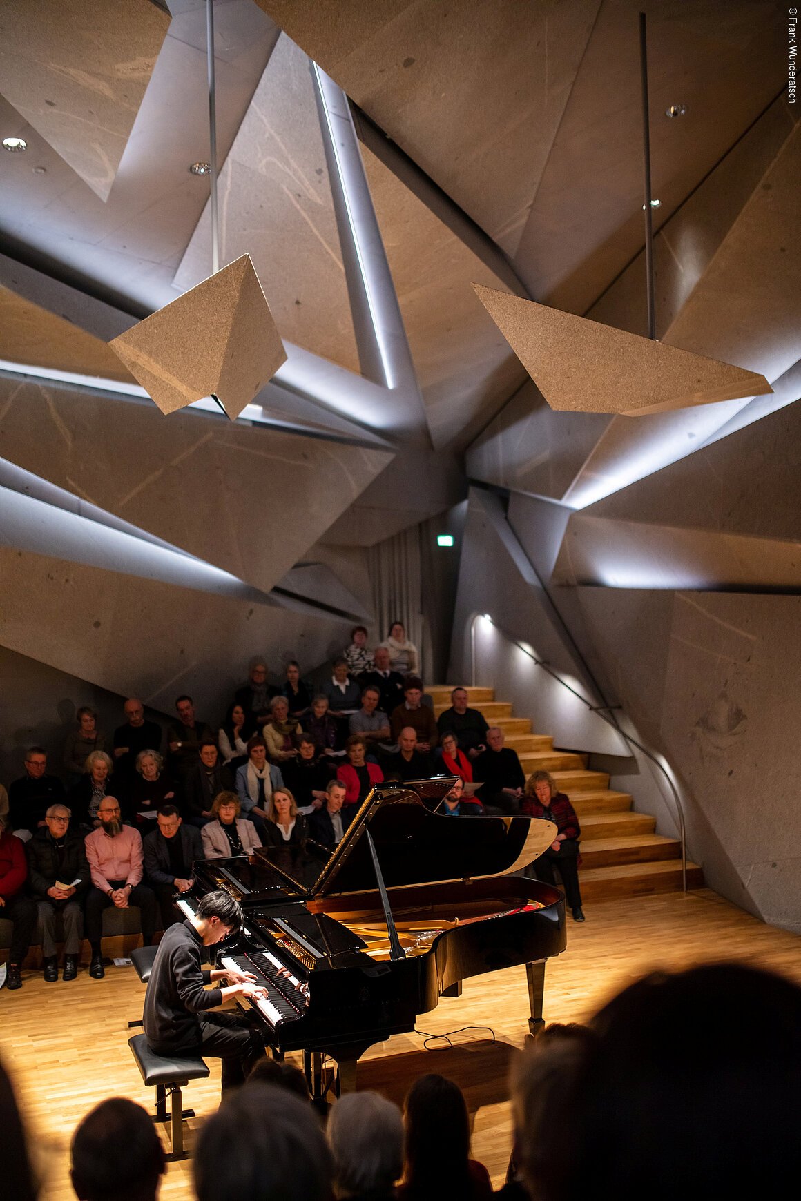 Eine Person sitzt an einem Flügel und spielt darauf. Im Hintergrund ist unscharf Publikum zu erkennen. Der Raum ist futuristisch mit ineinander geschachtelten riesigen Steinelementen.