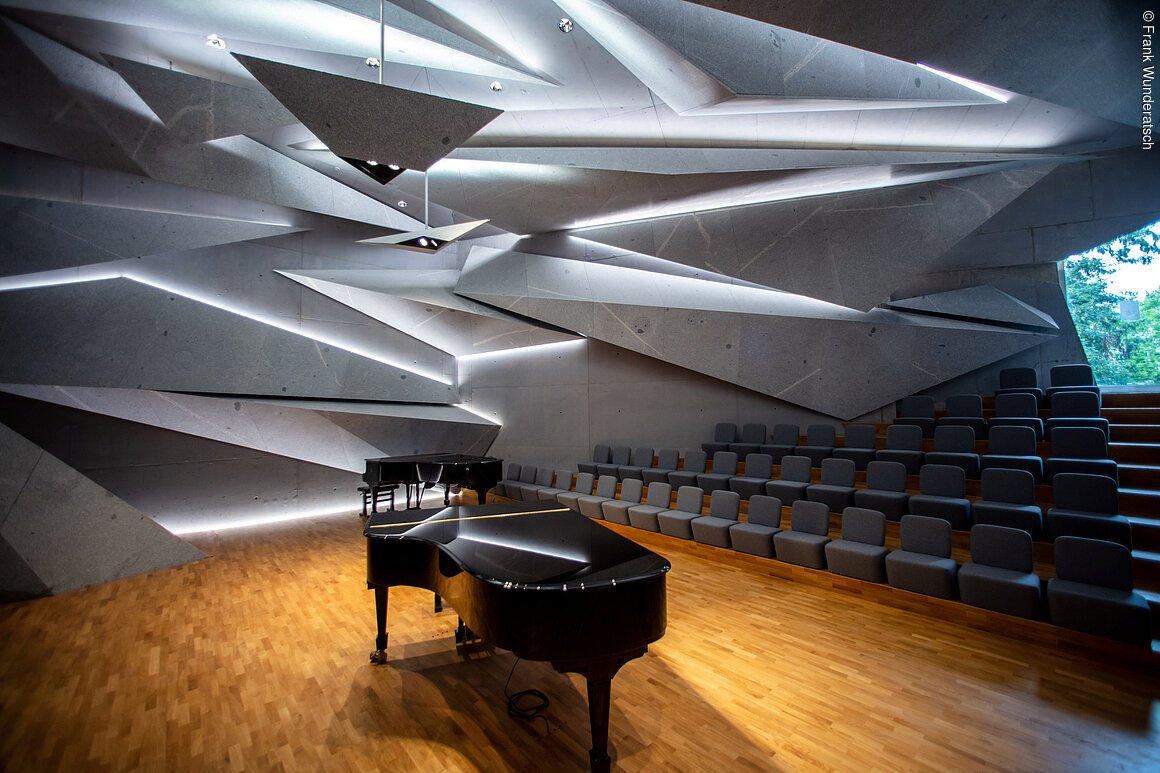 Zwei schwarze Konzertflügel mit Hocker stehen in einem mit Parkett ausgelegten großen Raum mit steinernen, spitz zulaufenden Bauelementen aus Stein, die geheimnisvoll beleuchtet und ineinander verschachtelt sind. Im Hintergrund sind Stuhlreihen zu erkennen.