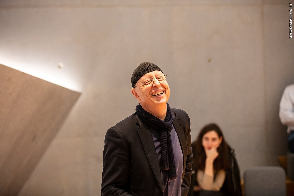 Ein Herr mit einer schwarzen Mütze steht vor einer Betonwand und lacht. Im Hintergrund ist unscharf eine sitzende junge Frau zu sehen.