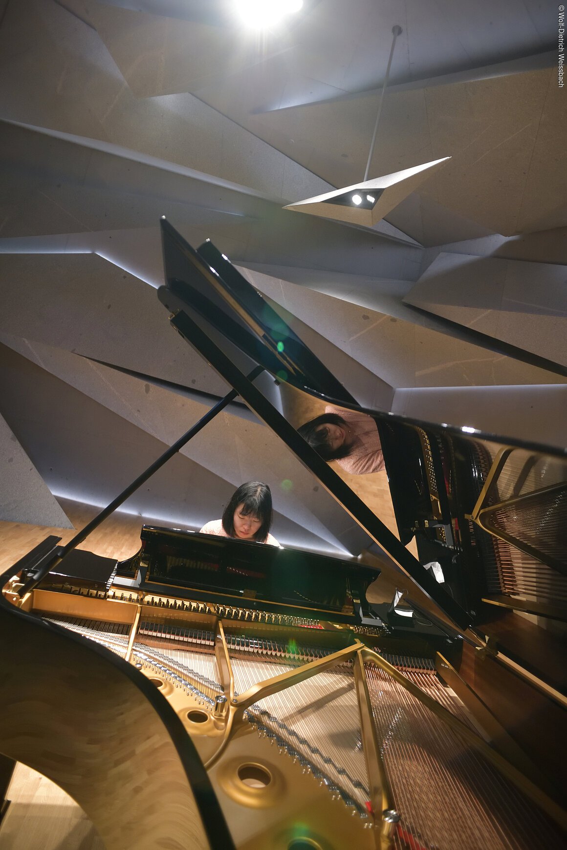 Eine Dame spielt an einem Flügel in einem modern gestalteten Raum, der aus Steinelementen besteht und geheimnisvoll beleuchtet ist..Der Blick geht durch den aufgestellten Flügel.