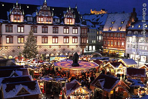 Ein Weihnachtsmarkt mit hell erleuchteten Buden, im nächtlichen Hintergrund eine ebenso illuminierte Burg
