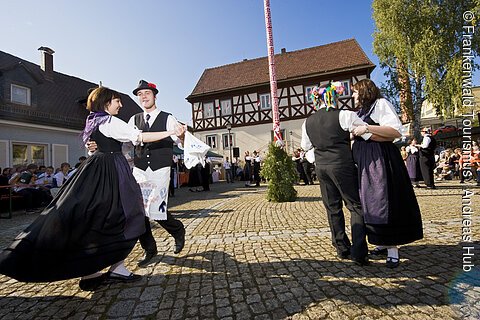 Tanzende Paare in traditioneller Tracht auf gepflastertem Platz vor Fachwerkhaus bei Fest mit Zuschauern.