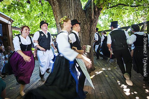 Menschen in traditioneller Tracht tanzen im Kreis auf einer Holzterrasse unter einem großen Baum.