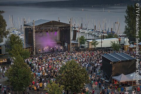 Open-Air-Konzert mit Bühne, Publikum und Segelbooten im Hafen im Hintergrund bei Abendlicht.