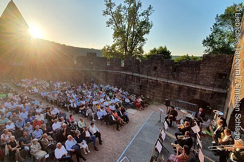 Publikum sitzt vor einer Freilichtbühne in einer Burgruine bei Sonnenuntergang, Musiker spielen Blasinstrumente.