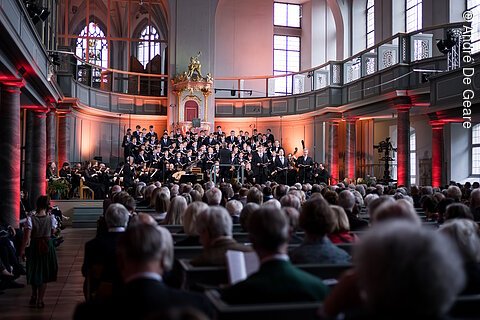 Kircheninnenraum mit Chor und Orchester vor Publikum bei Konzertveranstaltung mit roter Beleuchtung an Säulen