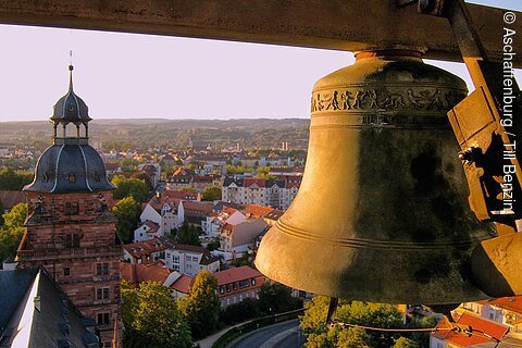 Große Kirchenglocke im Turm mit Blick auf Stadt, Häuser und Kirchturm im Hintergrund bei Sonnenuntergang.
