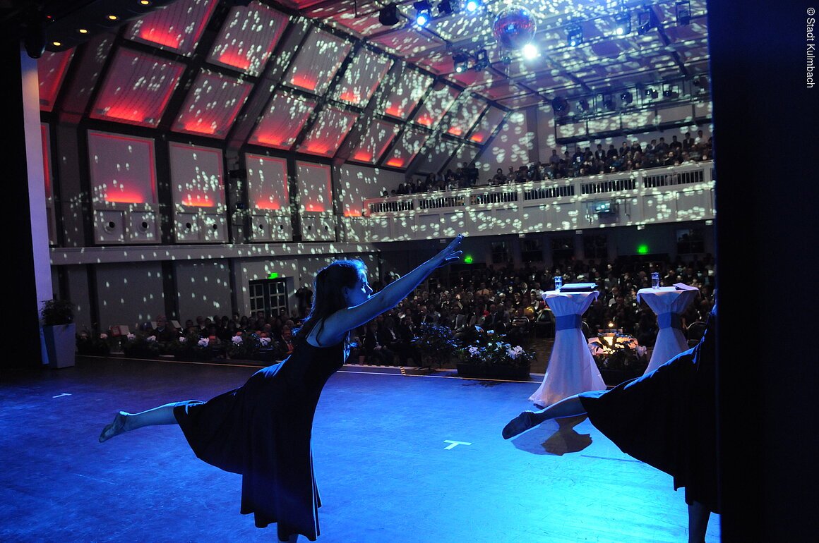 Tänzerin in schwarzem Kleid auf Bühne vor Publikum mit Disco-Lichteffekt an Decke und Wänden