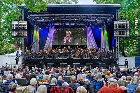 Orchester spielt auf Bühne im Freien vor Publikum, Dirigent auf großer Leinwand zu sehen, umgeben von Bäumen.