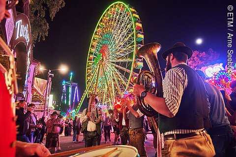Musiker in Trachten spielen Blasinstrumente vor beleuchtetem Riesenrad auf Nachtjahrmarkt mit Zuschauern.