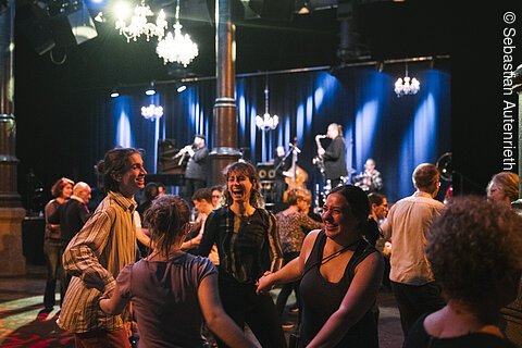 Menschen tanzen und lachen in einem Saal mit Live-Band und blauen Bühnenlichtern im Hintergrund.