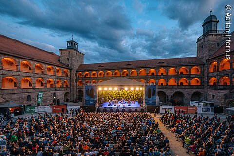 Große Menschenmenge vor beleuchteter Bühne im Innenhof eines historischen Gebäudes bei Abenddämmerung.