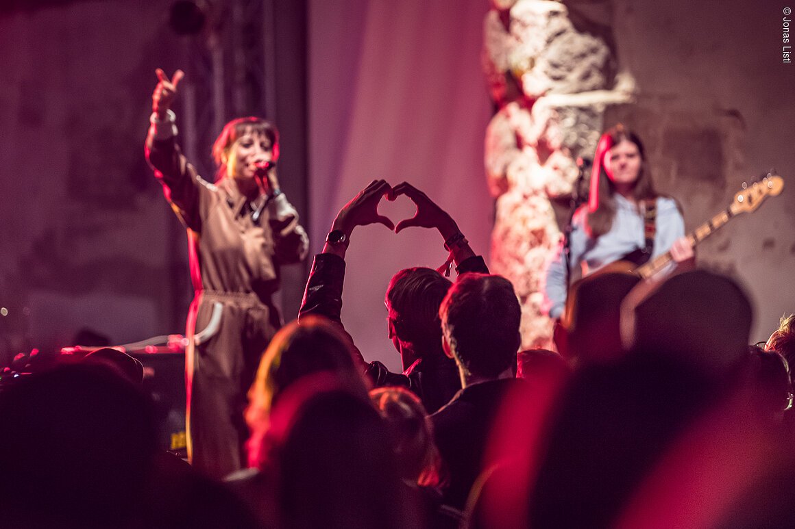 Publikum bei Konzert formt Herz mit Händen, Sängerin singt, Bassistin spielt Gitarre im Hintergrund.