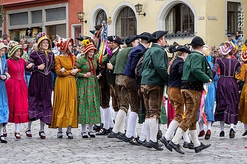 Menschen in historischer Tracht tanzen auf gepflastertem Platz vor Zuschauern und Gebäuden.