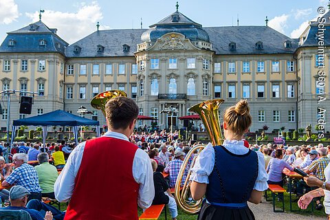 Zwei Musiker in Trachten mit Blasinstrumenten vor Publikum und großem Barockgebäude bei Tageslicht.