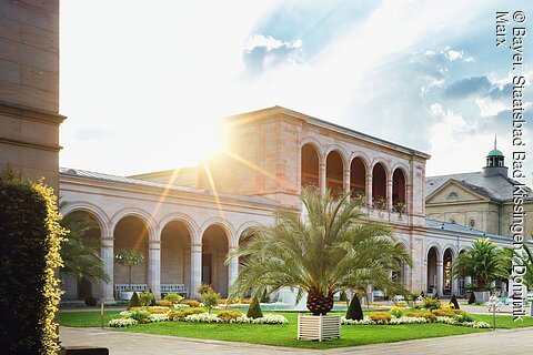 Sonniger Innenhof mit Palmen, Blumenbeeten und Arkadengang eines historischen Gebäudes.