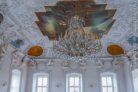 Eine Person in einem weißen Kleid geht durch einen prunkvollen Saal mit blauen Stühlen und einem großen Kronleuchter.
