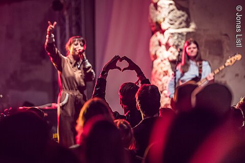 Publikum bei Konzert formt Herz mit Händen, Sängerin singt, Bassistin spielt Gitarre im Hintergrund.