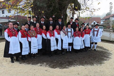 Gruppenfoto von Männern und Frauen in traditioneller Tracht vor großem Baum im Dorf.