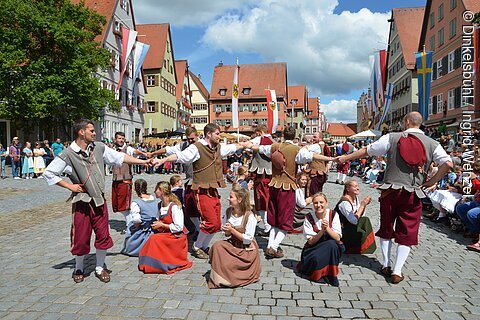 Traditionelle Trachten, Männer tanzen stehend, Frauen sitzen klatschend auf Kopfsteinpflaster in historischem Stadtbild.