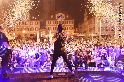 Blick von der Bühne auf ein Publikum bei einem nächtlichen Open-Air-Konzert mit Konfetti und Musikern.