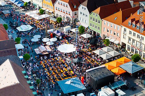 Vogelperspektive auf belebten Marktplatz mit Menschen, Tischen, Sonnenschirmen und bunten Häusern im Hintergrund.