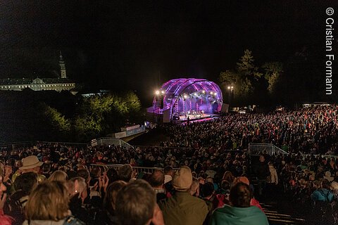 Große Menschenmenge bei Nacht vor beleuchtetem Konzertbühnenzelt im Freien mit Bäumen im Hintergrund.